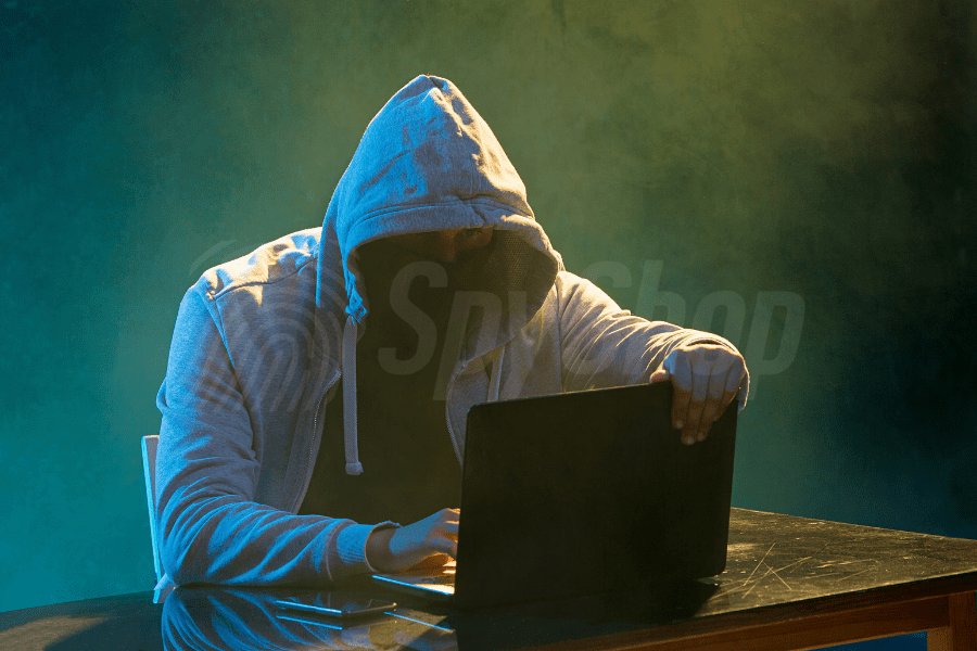 A masked man in a hooded sweatshirt sits at a table in a dark room. He is using a laptop.