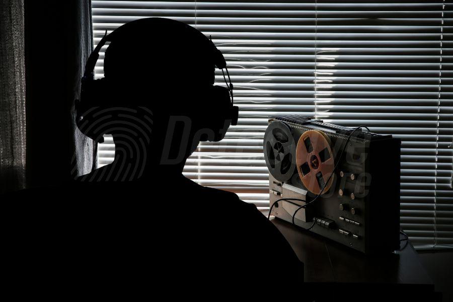 Silhouetted surveillance operator monitoring audio with a reel-to-reel recorder — illustrating the covert nature of laser microphone operations.