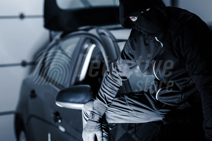 A masked man in black clothes and gloves, presumably a thief, is standing with one leg up on the doorstep of an open car in a dark garage.