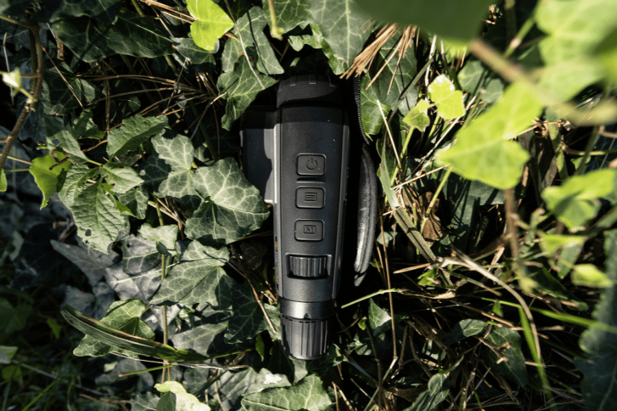 A monocular lying in the grass, partially hidden among ivy leaves. The device shows control buttons and a dial, with green plants and grass in the background.