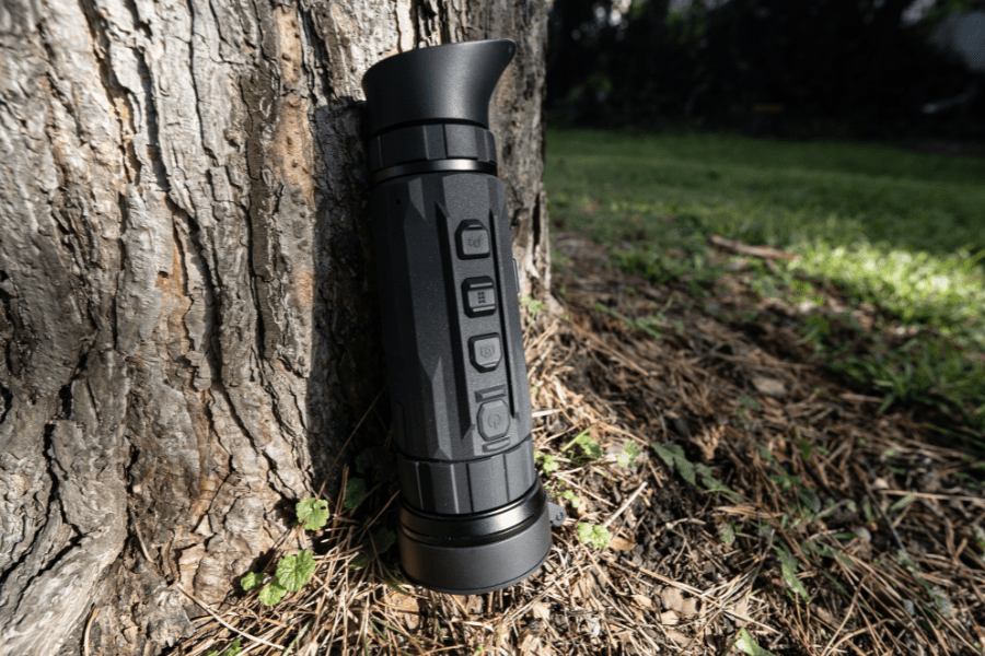 A monocular resting against a tree trunk in a forest, surrounded by grass and fallen leaves. Greenery can be seen in the background, and the control buttons are clearly visible on the device.