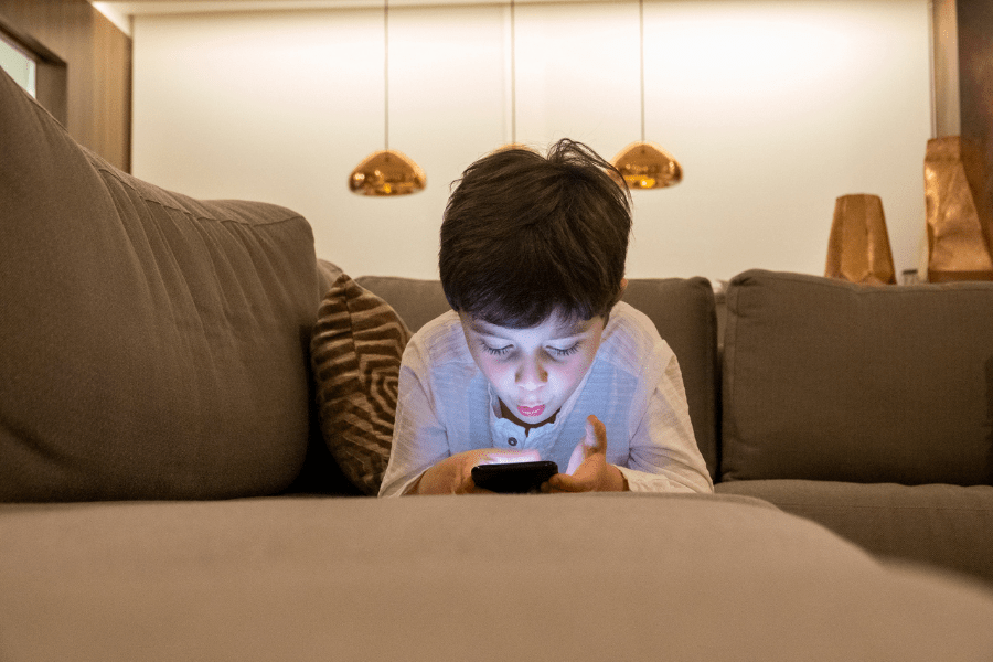 A little boy is lying on the corner of the sofa, talking on the phone, a blue glow reflecting on his face. In the background, there is living room furniture.