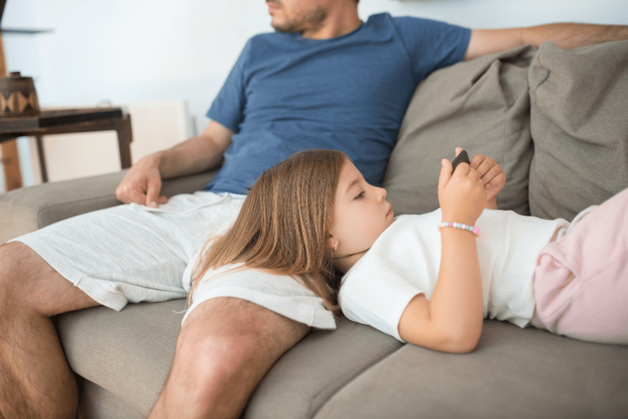 A girl is lying on her dad's lap and browsing through her phone.