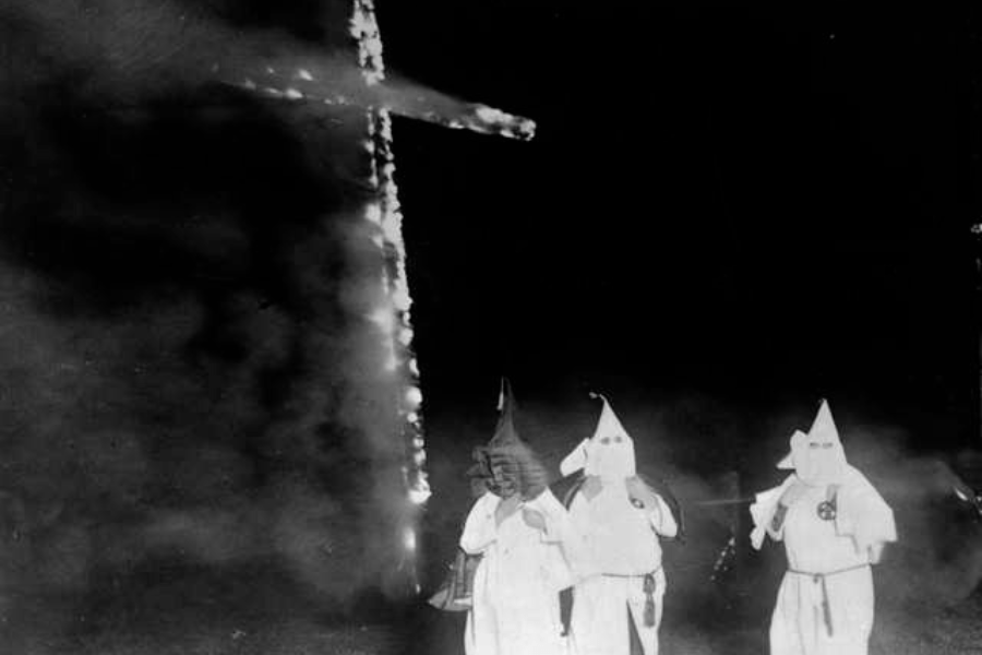 صورة بالأبيض والأسود تُظهر ثلاثة من أعضاء كو كلوكس كلان بزيّهم الرسمي، مع صليب مشتعل في المقدمة.